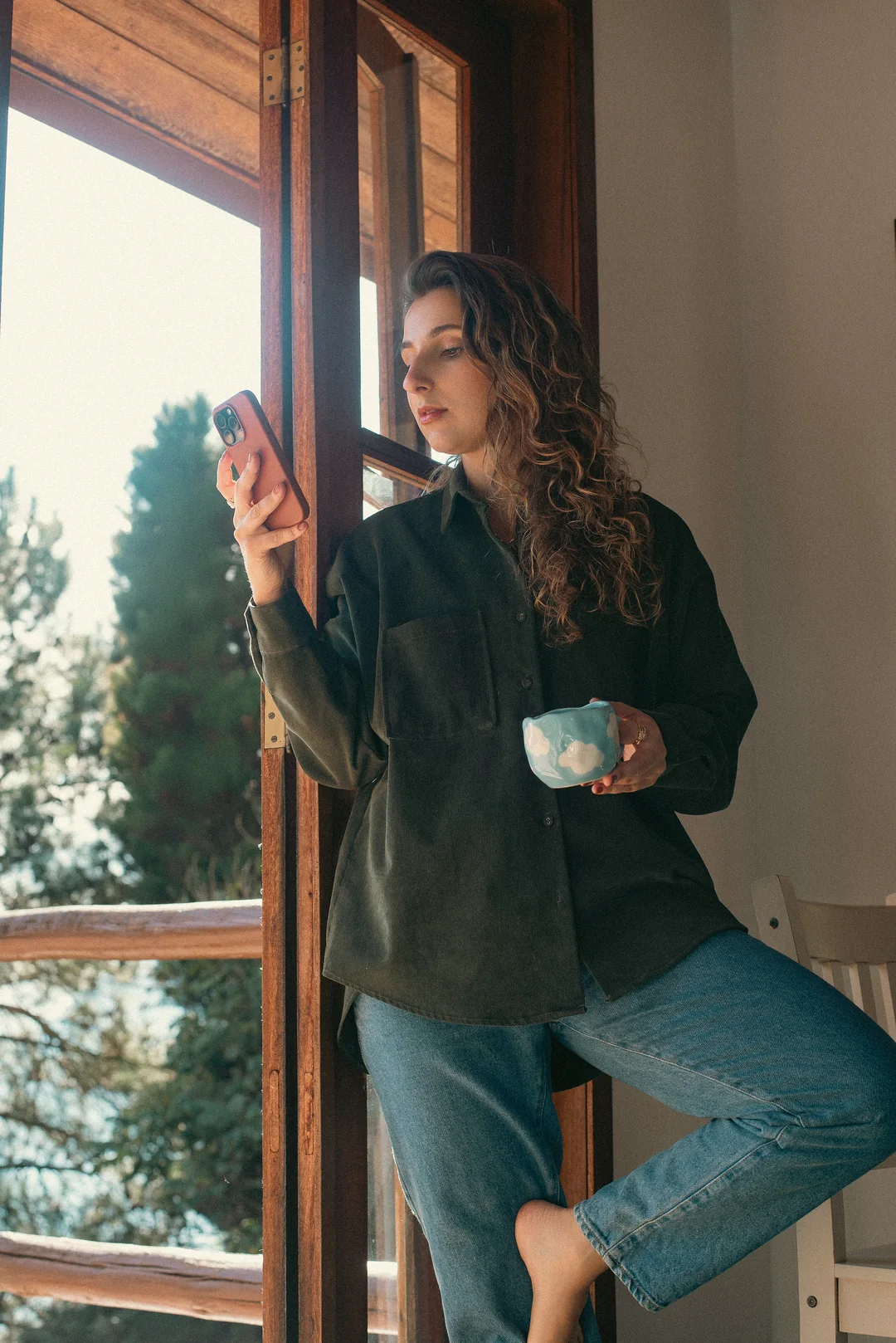 Isabela Vicarpi na janela, com o celular numa mão e uma caneca na outra